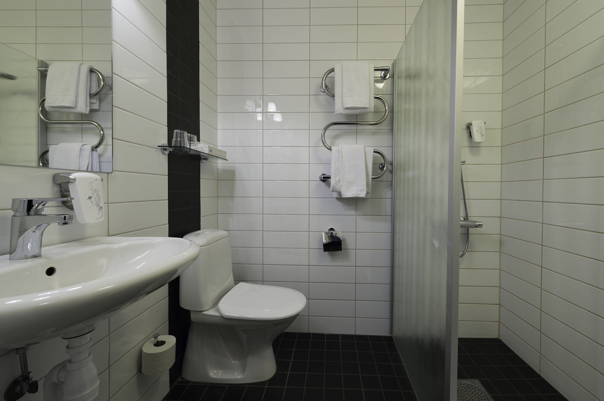 Un baño con lavamanos, inodoro y ducha blancos. Hay toallas colgadas en toalleros y un dispensador de papel higiénico.