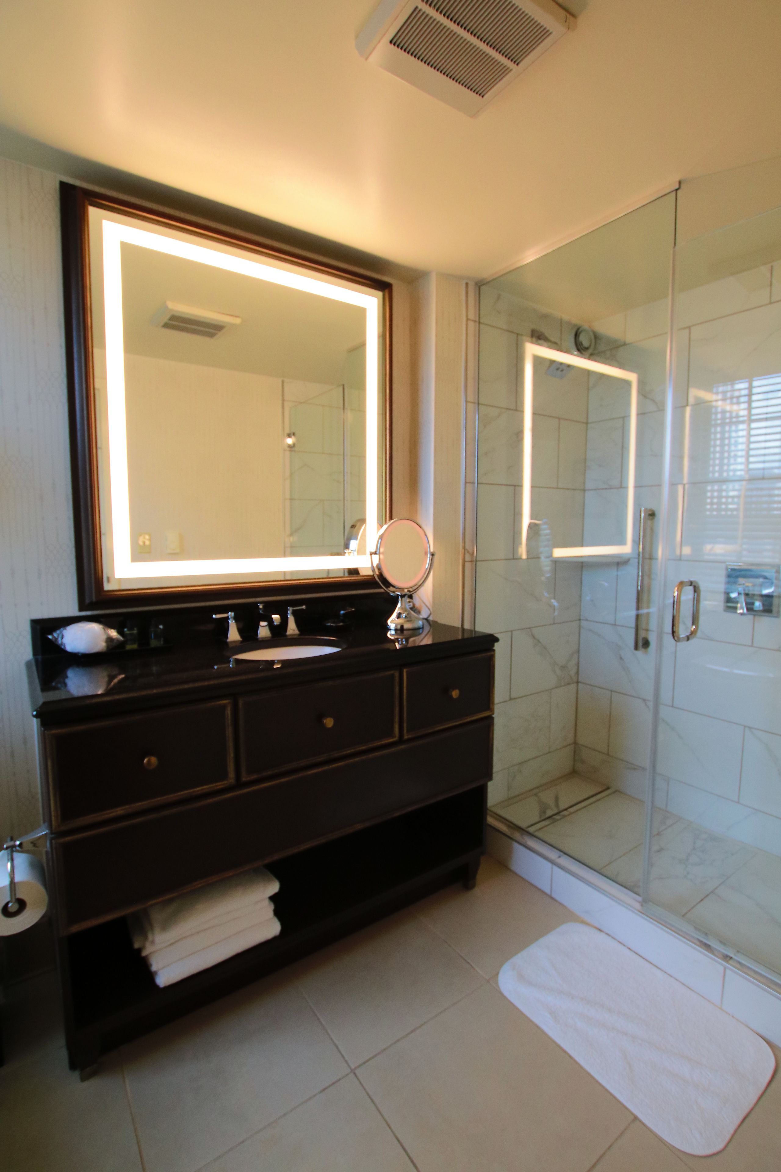 A bathroom with a large mirror, a sink, a vanity, a glass shower enclosure, and a white bath mat.