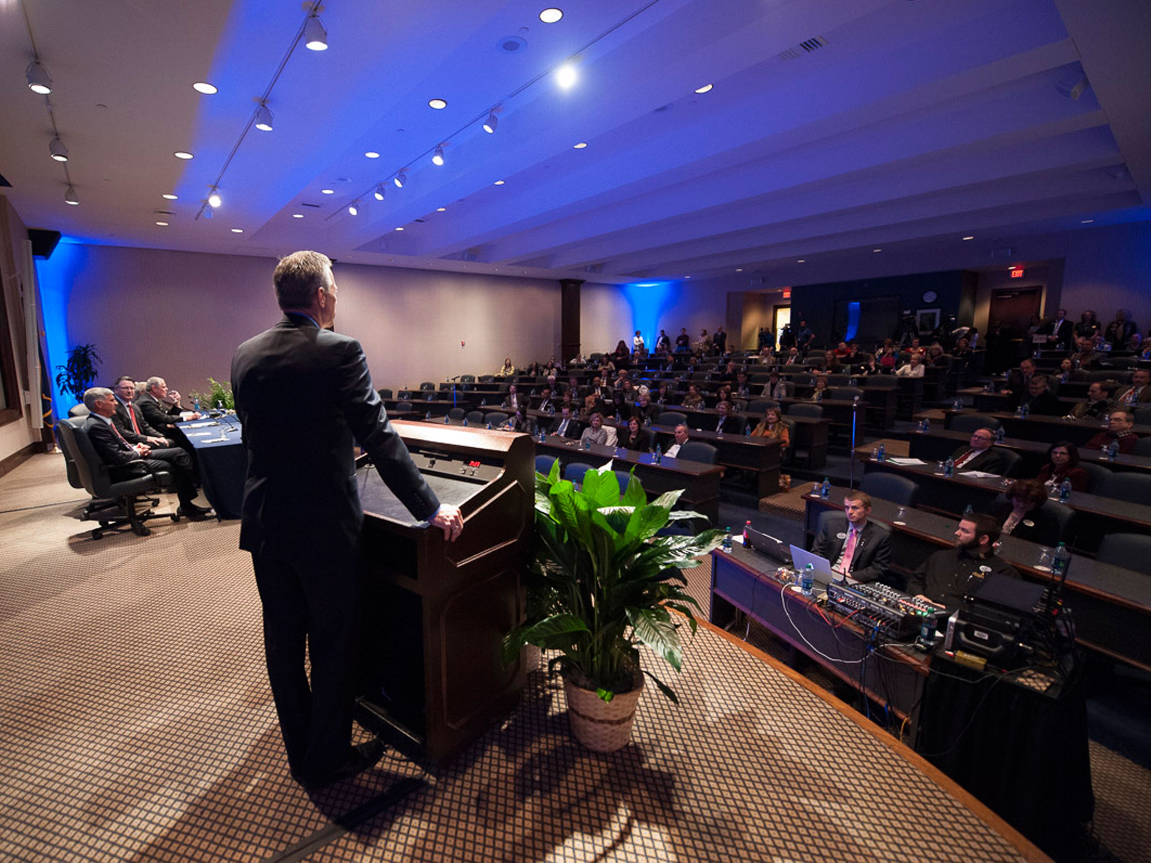 A conference room with a speaker at a podium, attendees seated at tables, and a stage with a plant.