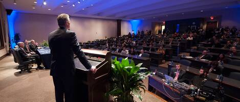 A conference room with a speaker at a podium, attendees seated at tables, and a stage with a plant.