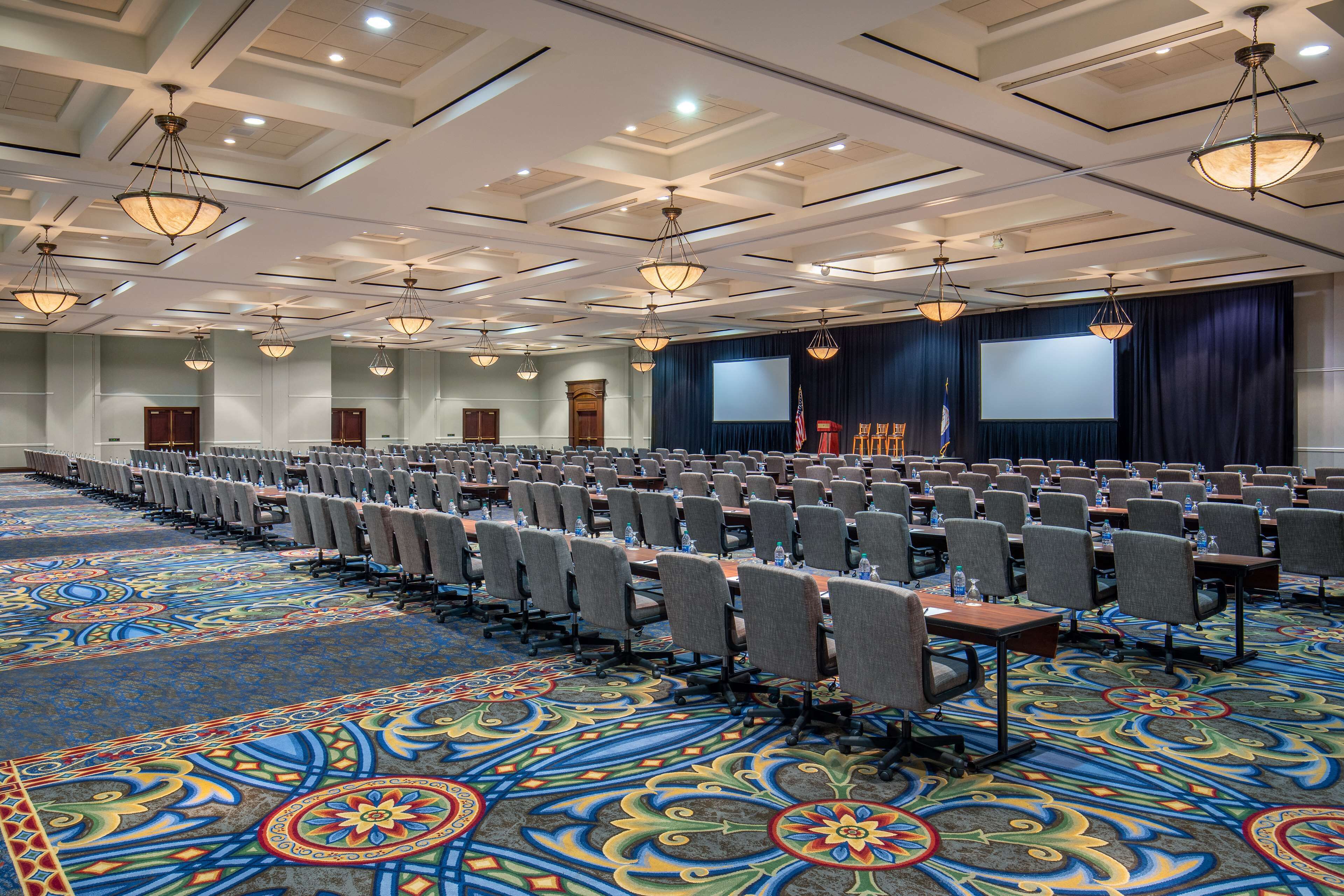A large conference room with rows of tables and chairs, a patterned carpet, and a stage with a screen.