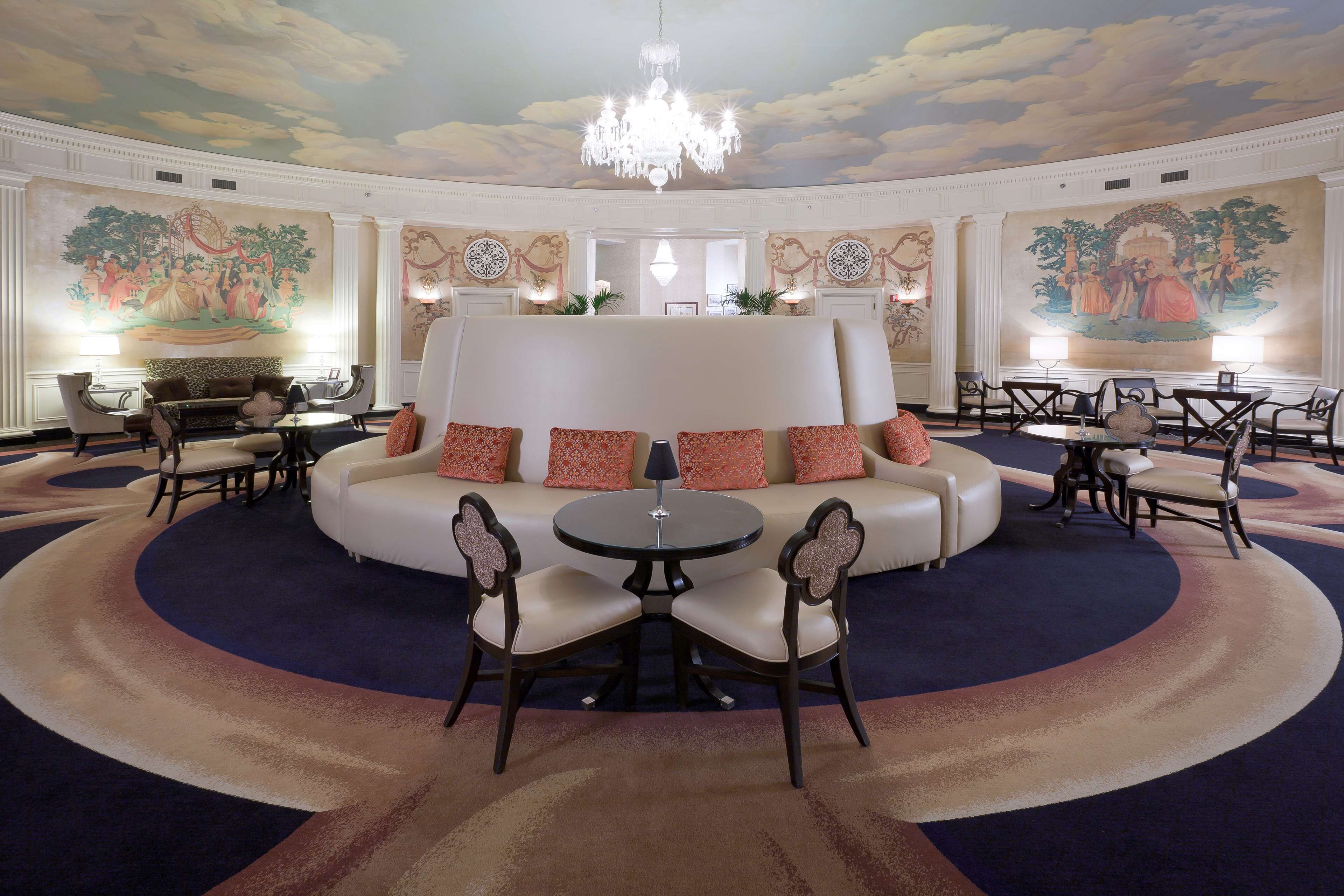 A luxurious hotel lobby with a central seating area, ornate wall murals, and a grand chandelier.