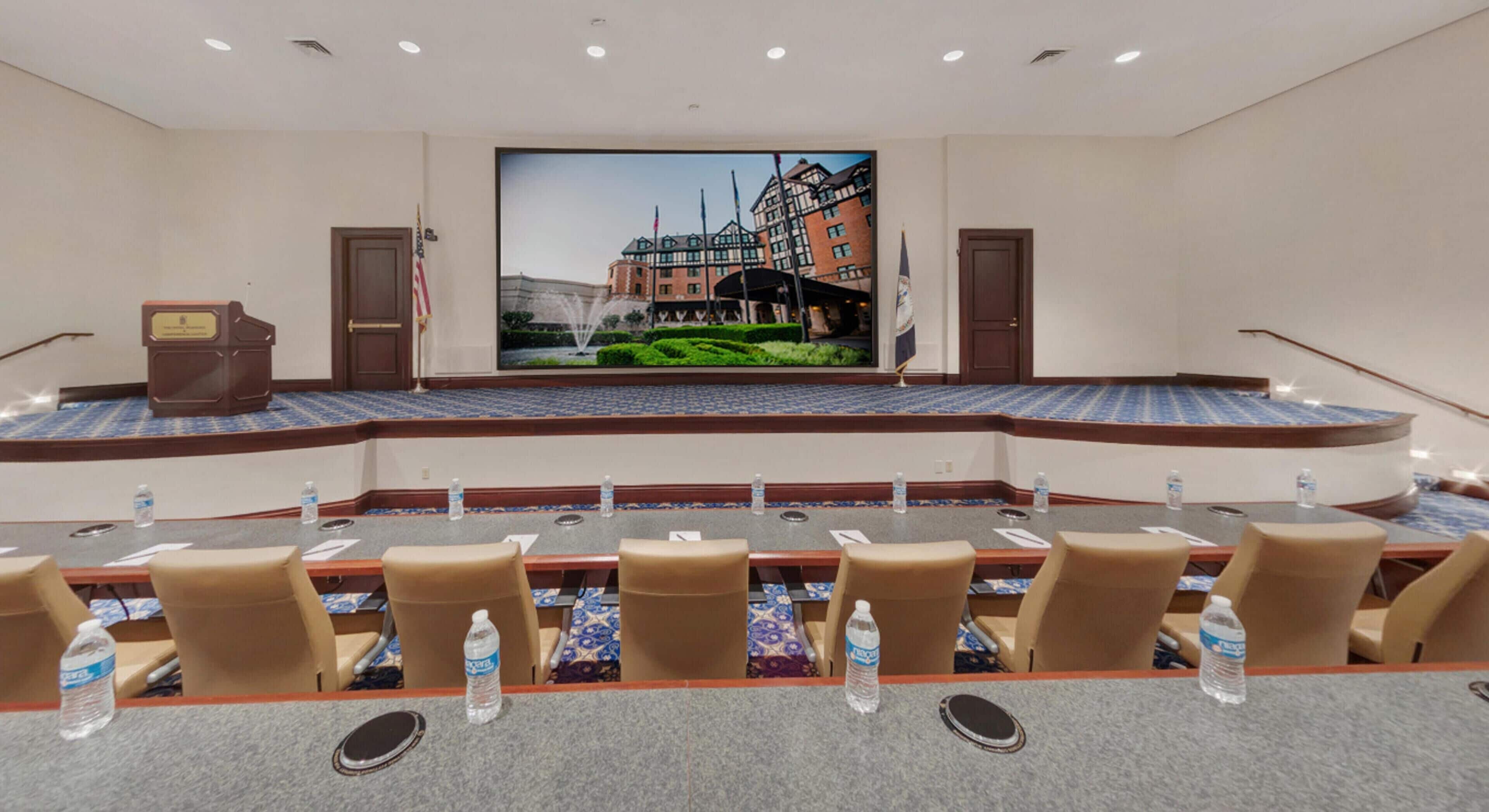 Una sala conferenze con un grande schermo che mostra un edificio, un leggio e bottiglie d'acqua sul tavolo.