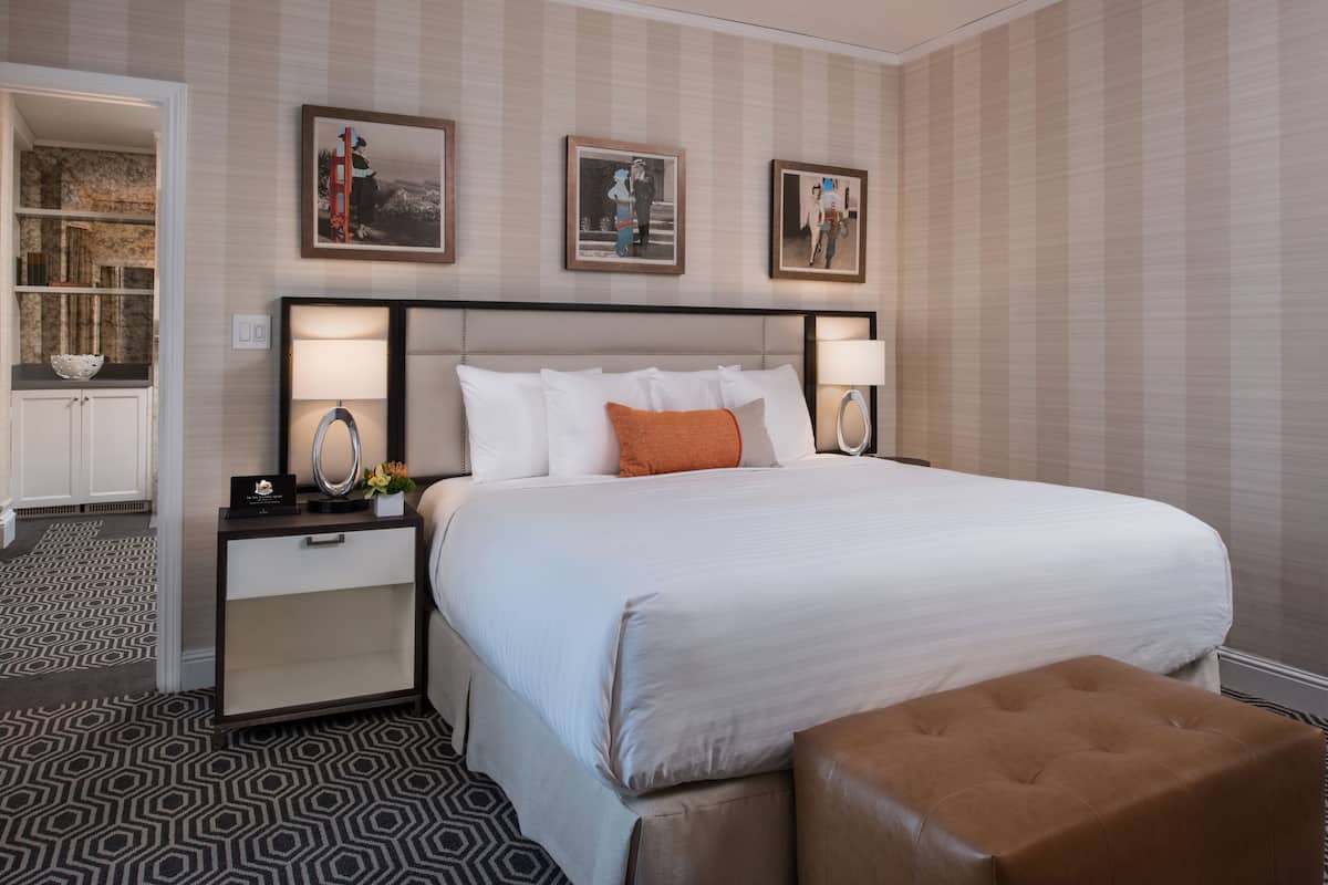 A hotel room with a large bed, two bedside tables with lamps, a small side table, a decorative bowl, and a framed picture above the bed.