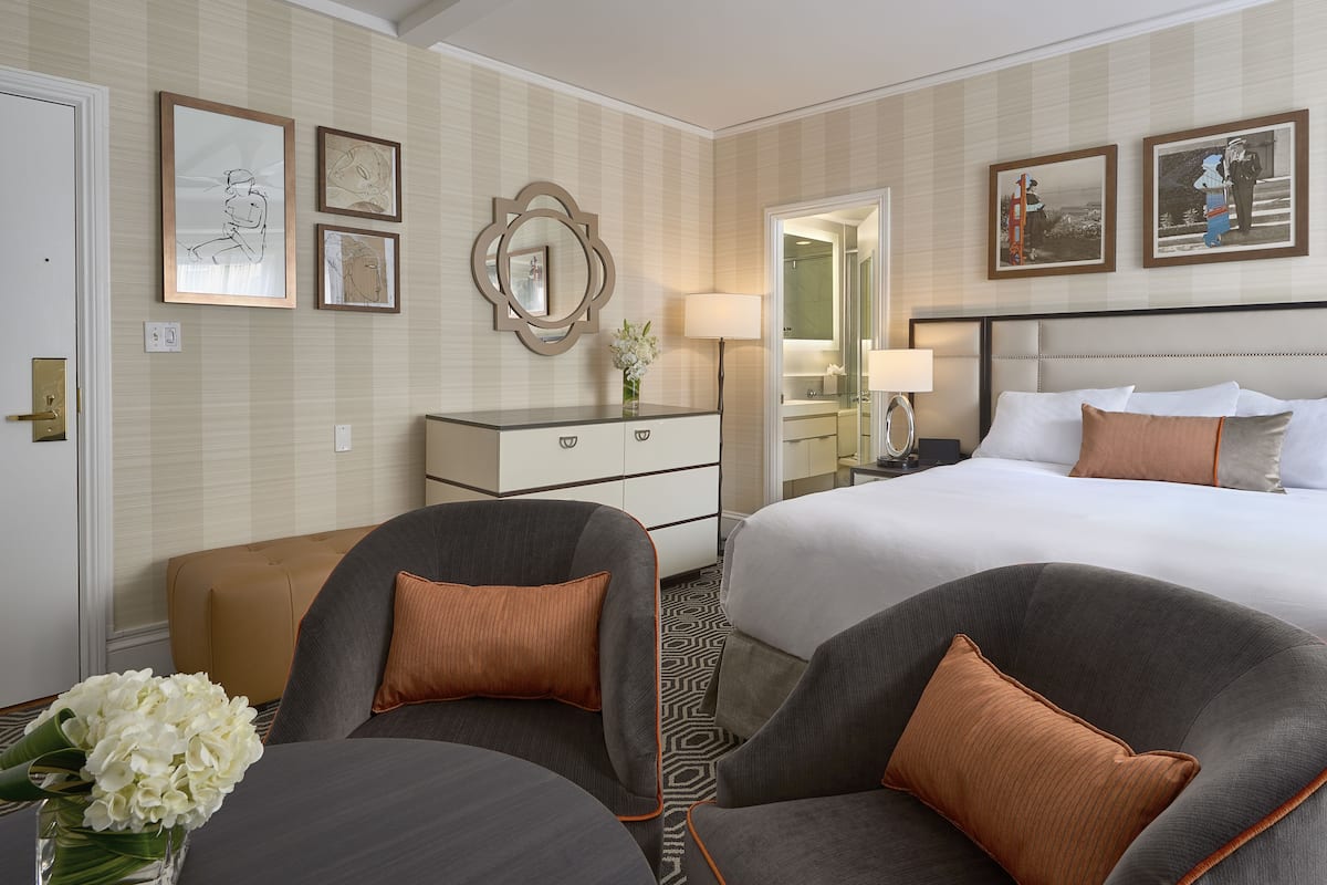 A hotel room with a bed, two armchairs, a nightstand, a dresser, and framed artwork on the walls.