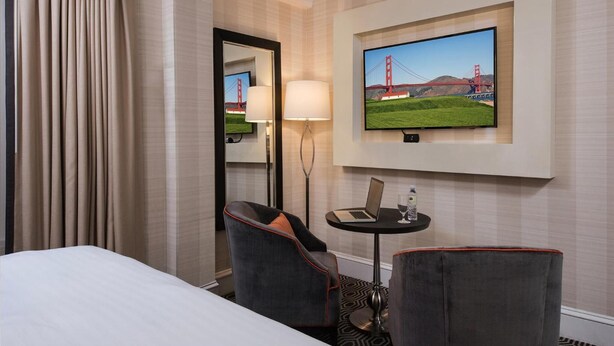 A hotel room with a bed, two armchairs, a small table with a laptop and a glass of water, a lamp, a TV displaying a scenic image, and a large window with curtains. - Inn At Union Square (San Francisco)