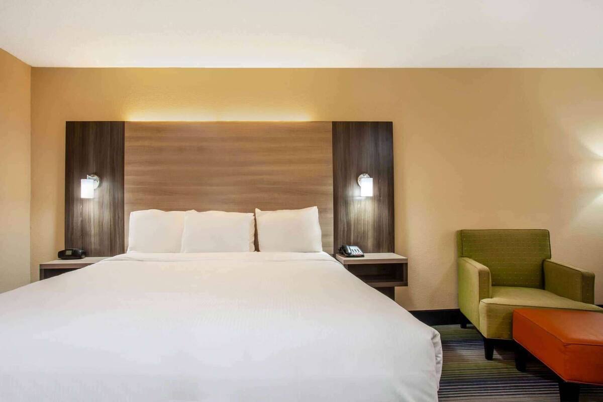 Ein Hotelzimmer mit einem großen Bett, zwei Nachttischen mit Lampen, einem grünen Sessel und einem kleinen orangefarbenen Pouf.