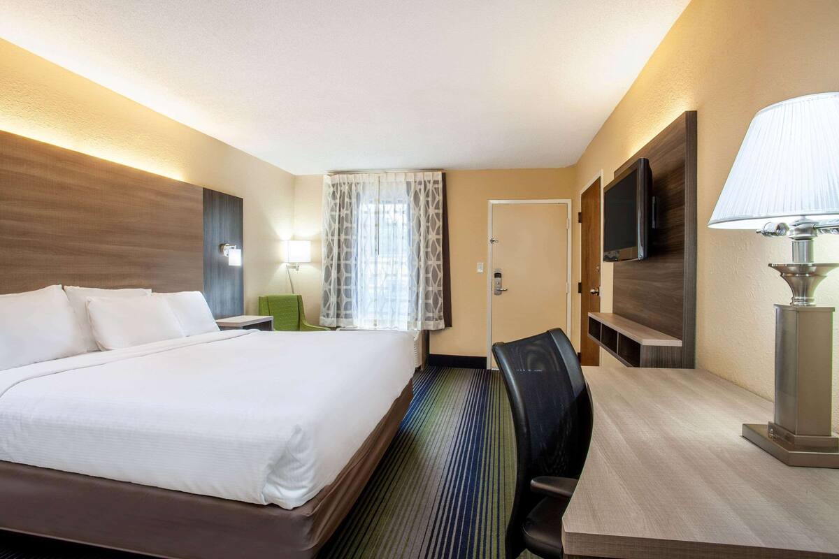 Ein Hotelzimmer mit einem großen Bett, einem Schreibtisch mit Lampe, einem Sessel, einem Fernseher und einem Fenster mit Vorhängen.