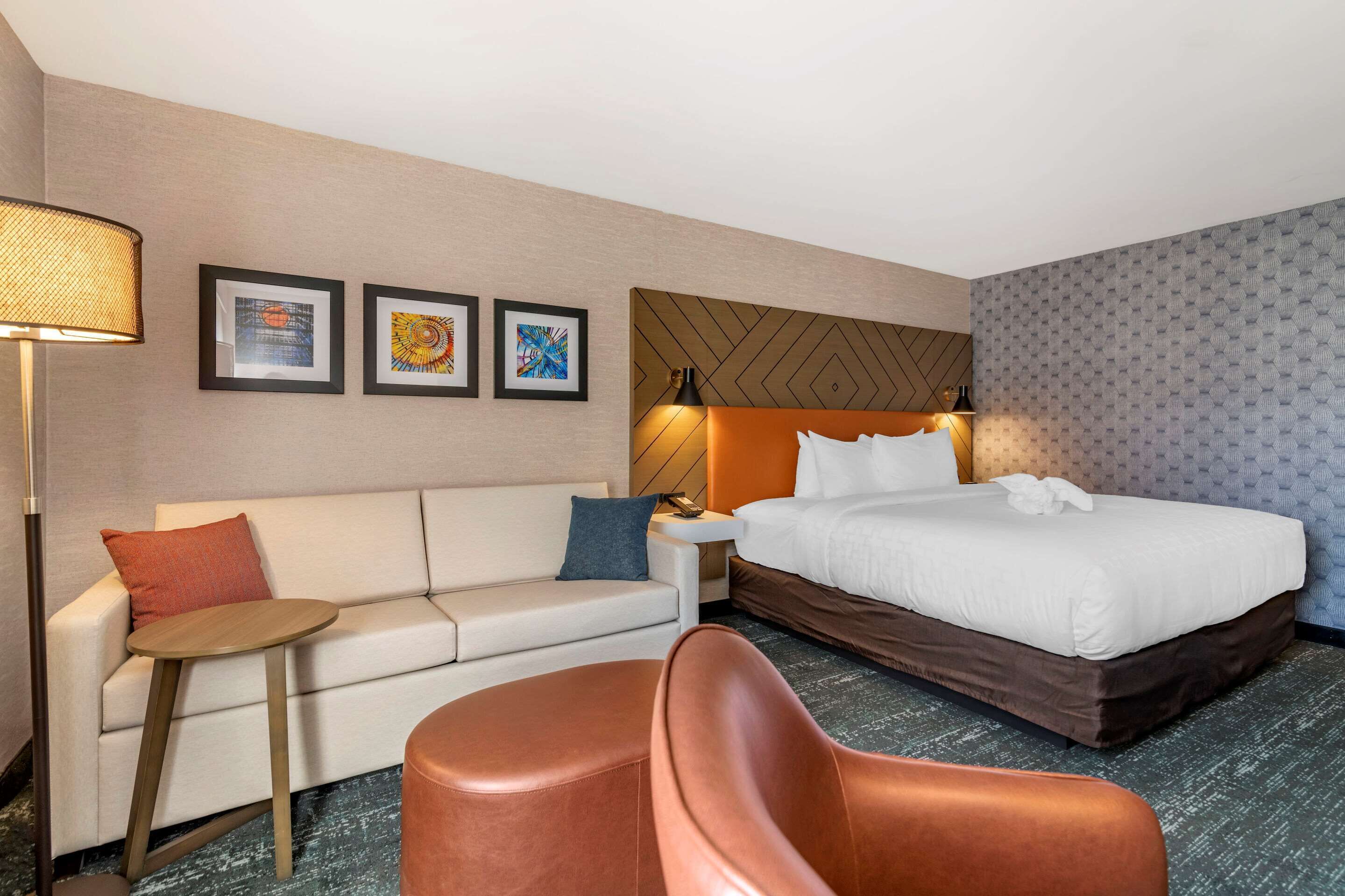 A hotel room with a bed, a sofa, a small table, and a chair. There are three framed pictures on the wall.