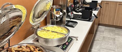 Montaje de bufé con diferentes alimentos, como huevos revueltos, salchichas y un dispensador de sirope de arce.