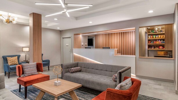 A modern lobby with a grey sofa, red armchairs, a wooden coffee table, and a reception desk with a wooden panel design. - Clarion Inn Nashua (Nashua)