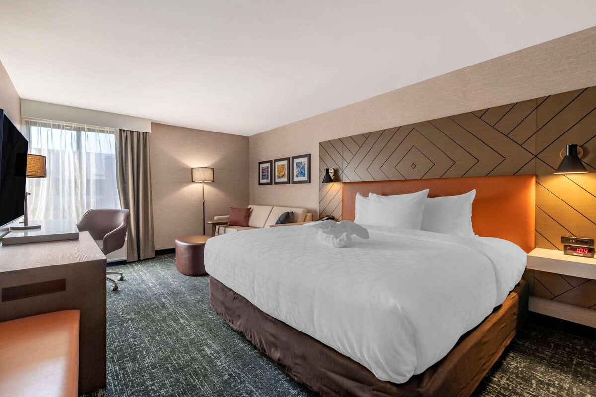 Habitación de hotel con una cama grande, un escritorio, una silla, un televisor y un reloj en la mesita de noche.