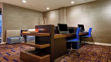 A modern hotel room with a wooden desk, two blue office chairs, a laptop, and a small seating area with a sofa and ottoman.