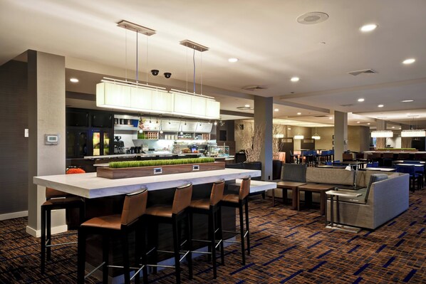 A modern hotel lobby with a central bar area, seating arrangements, and a kitchen in the background. - Courtyard By Marriott Cape Cod Hyannis (Hyannis)