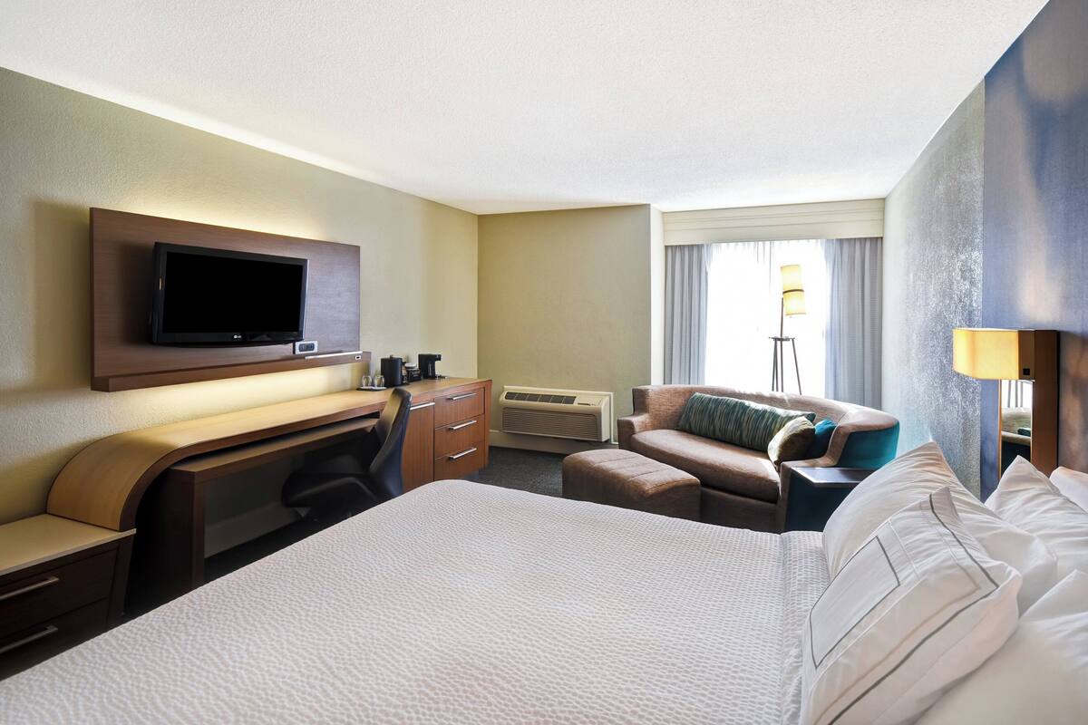 A hotel room with a large bed, a desk with a flat-screen TV, a sofa, and a lamp.