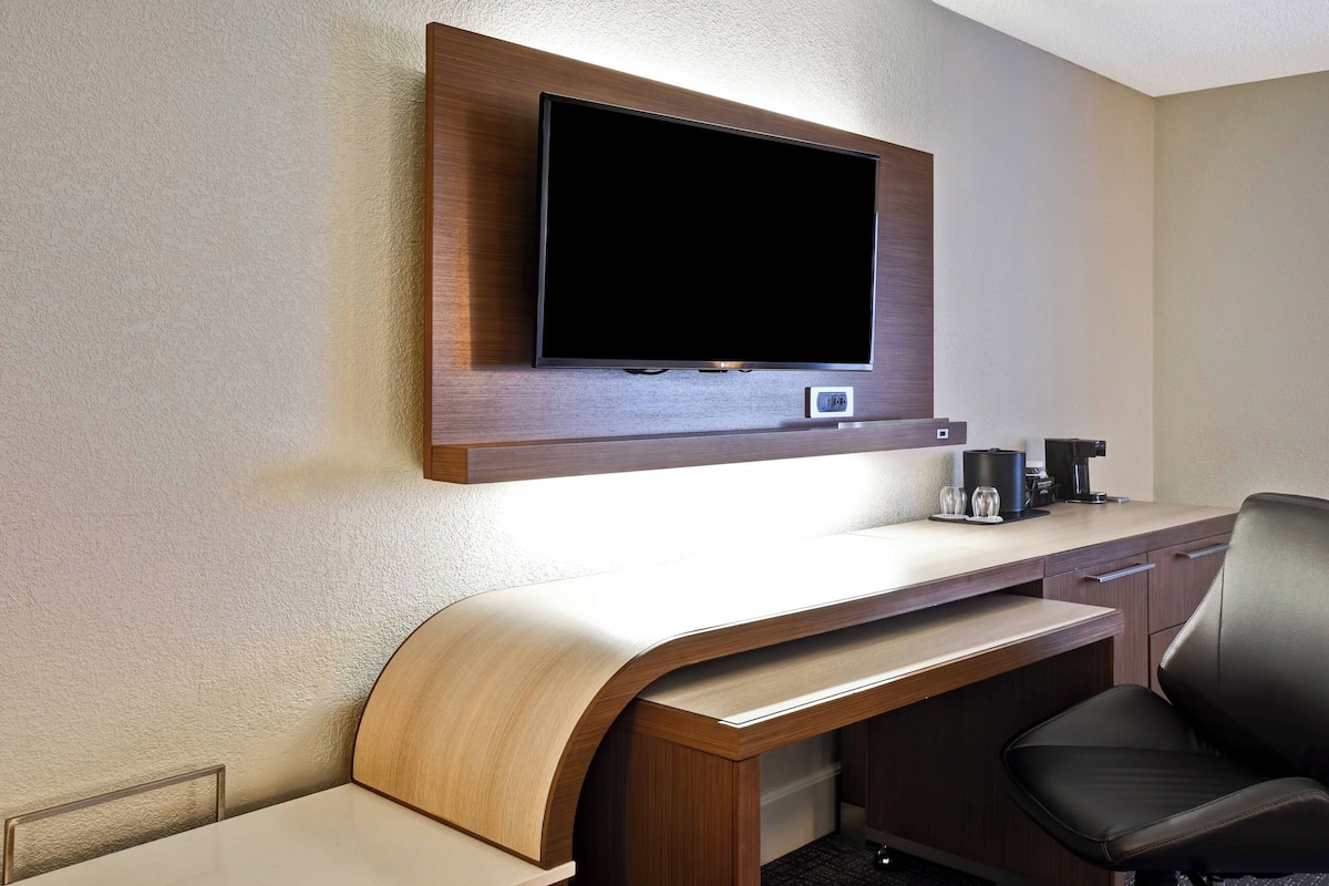 A hotel room with a flat-screen TV mounted on the wall, a wooden desk with a curved front, a black leather chair, and a coffee maker on the desk.