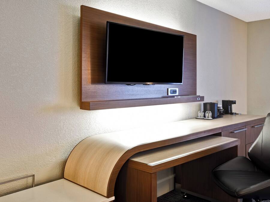 Een hotelkamer met een flatscreen televisie aan de muur, een houten bureau met een gebogen voorkant, een zwarte lederen fauteuil en een koffiezetapparaat op het bureau.