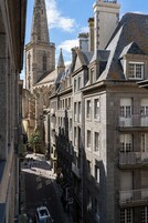 Una estrecha calle bordeada por edificios históricos de piedra, con varios pisos, amplias ventanas y una imponente torre de iglesia.