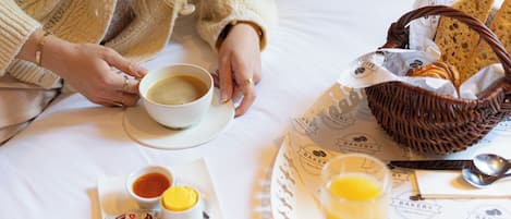 A breakfast setup on a bed with a cup of coffee, a plate with Nutella and jam, a glass of orange juice, and a basket of bread and pastries.