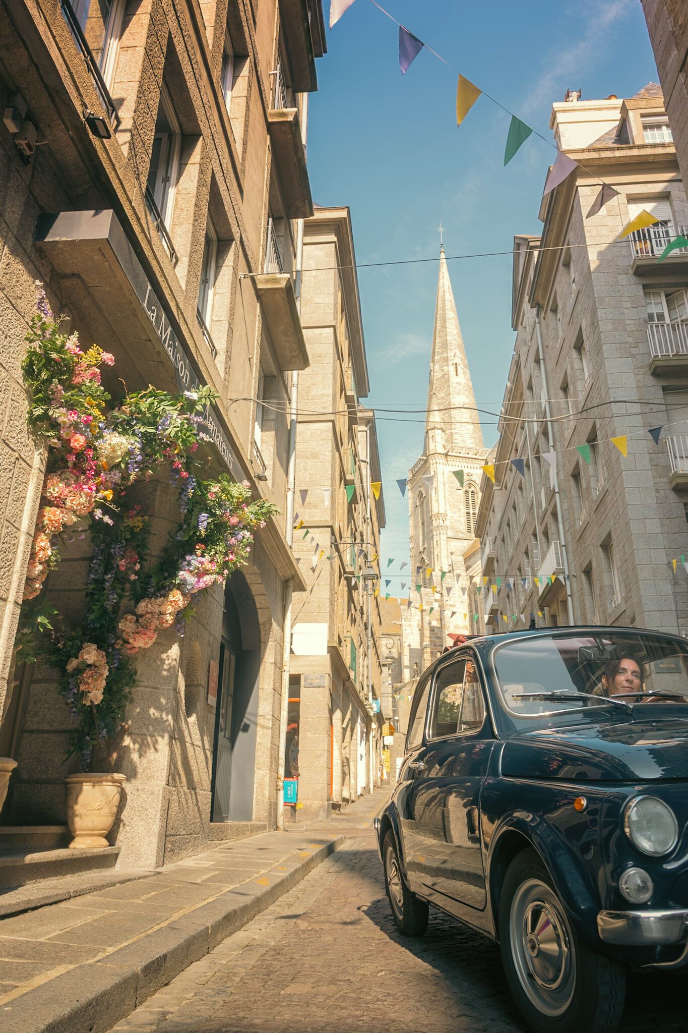 Una calle estrecha con edificios históricos, un coche clásico y banderas coloridas.