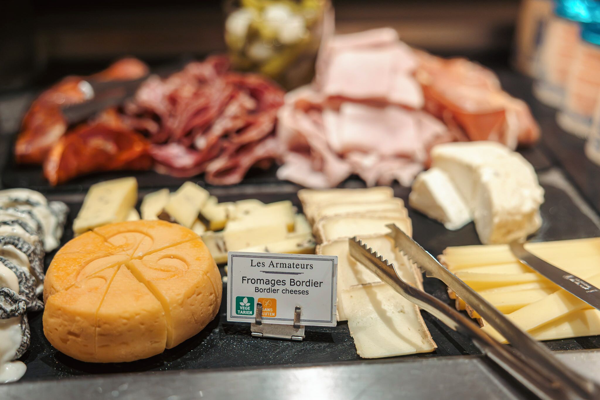 Una tabla de quesos con diferentes tipos, incluyendo un queso redondo con un logotipo, y un cartel que indica 'Fromages Bordier'.