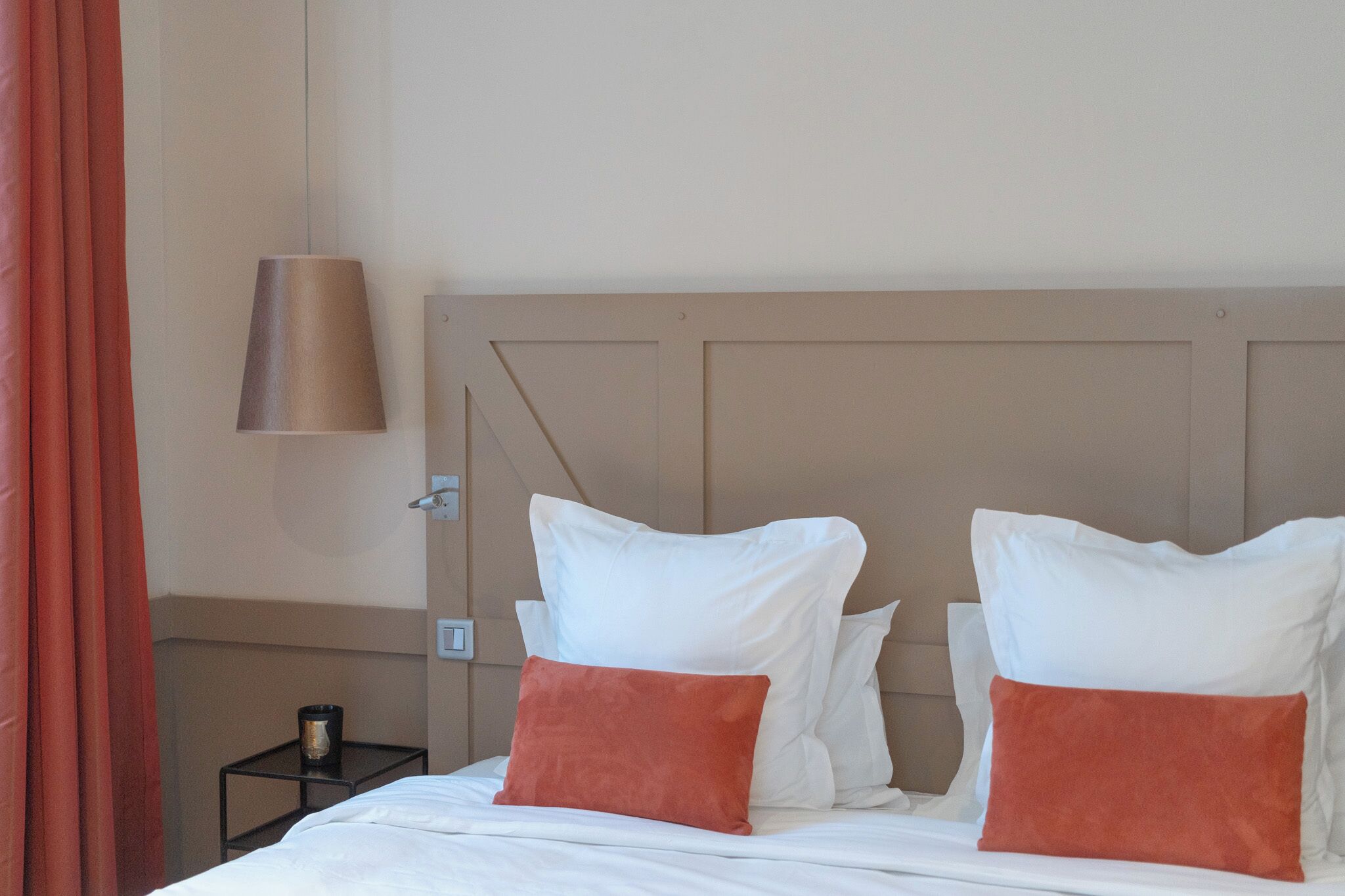 A hotel room with a bed, two white pillows, one orange pillow, a bedside table with a lamp, and a red curtain.