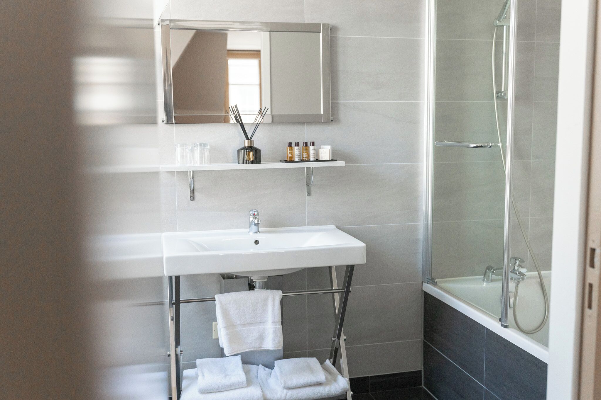 A modern bathroom with a white sink, mirror, and shower. There are towels and toiletries on the sink.