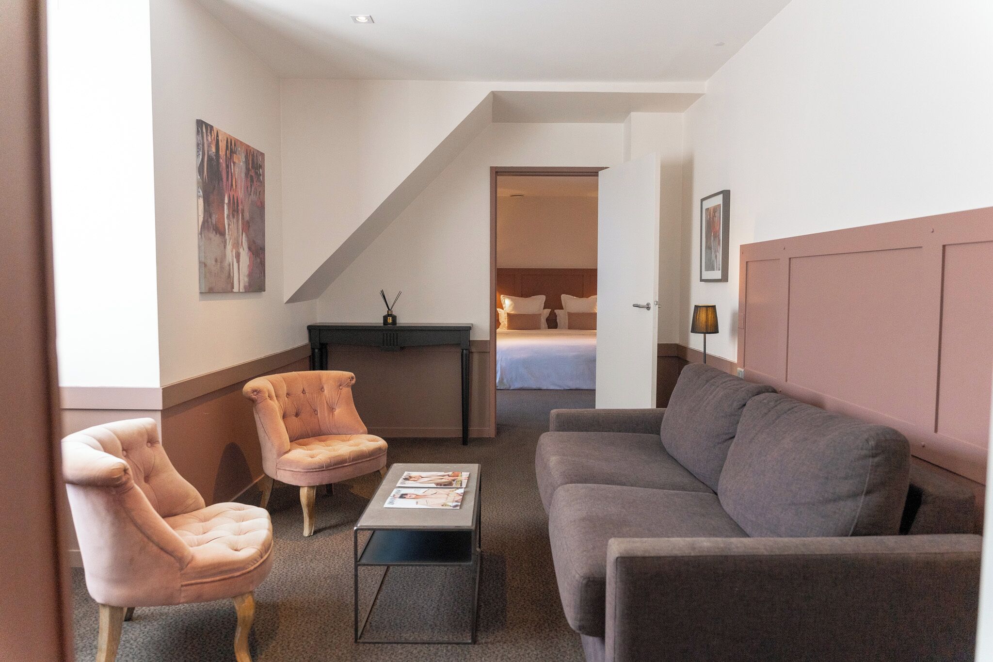 A hotel room with a grey sofa, two armchairs, a small table with a magazine, a desk with a lamp, and a bed in the background.