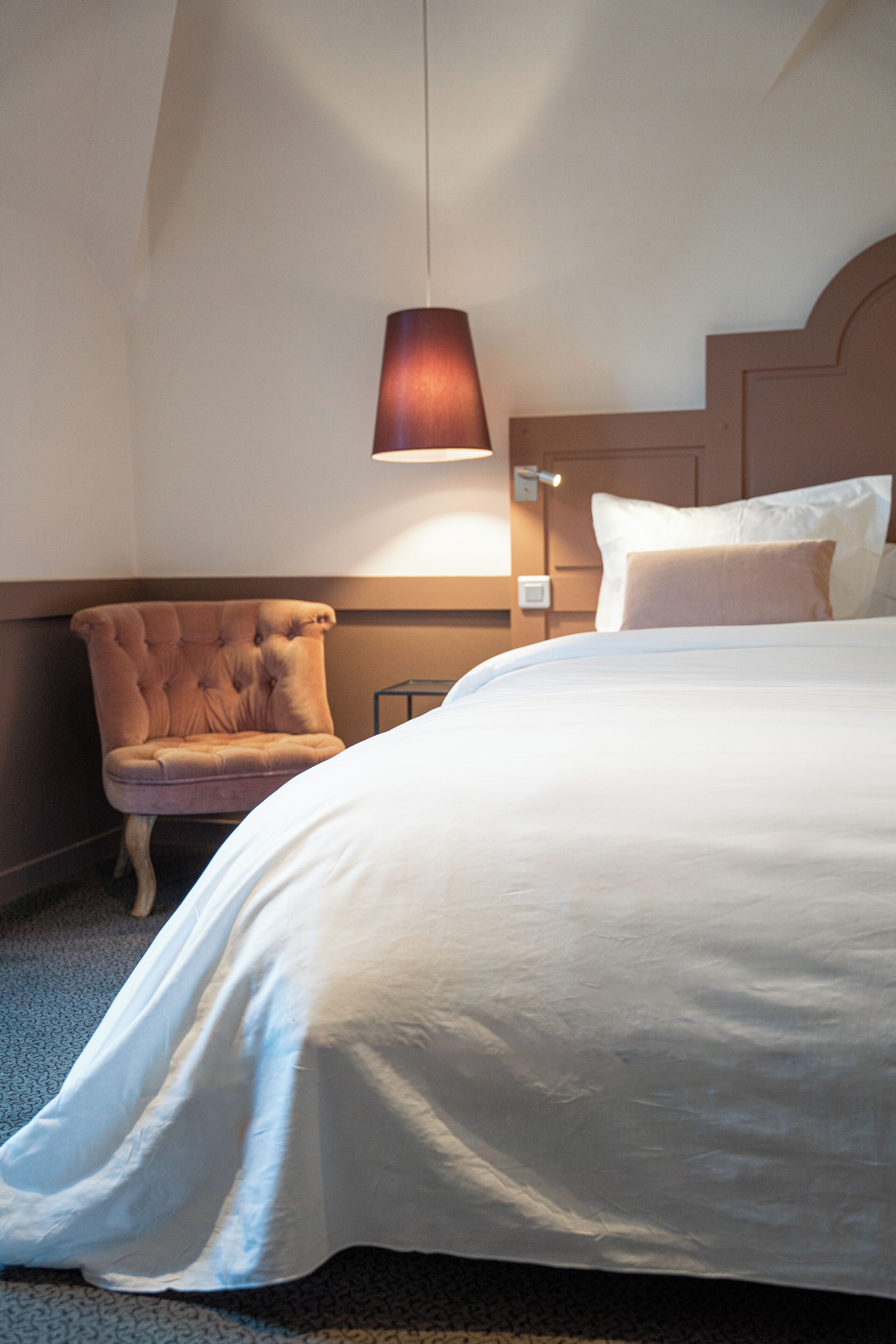 Una habitación de hotel con una cama grande, un sillón capitonado, una lámpara de pared y una mesita de noche.