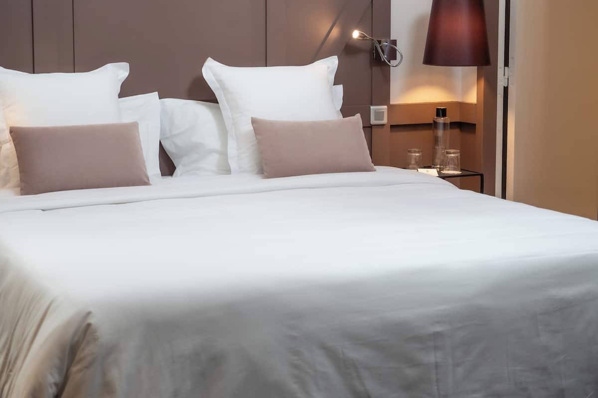A neatly made bed with white and beige bedding, a wall-mounted lamp, and a small bedside table with a glass container.