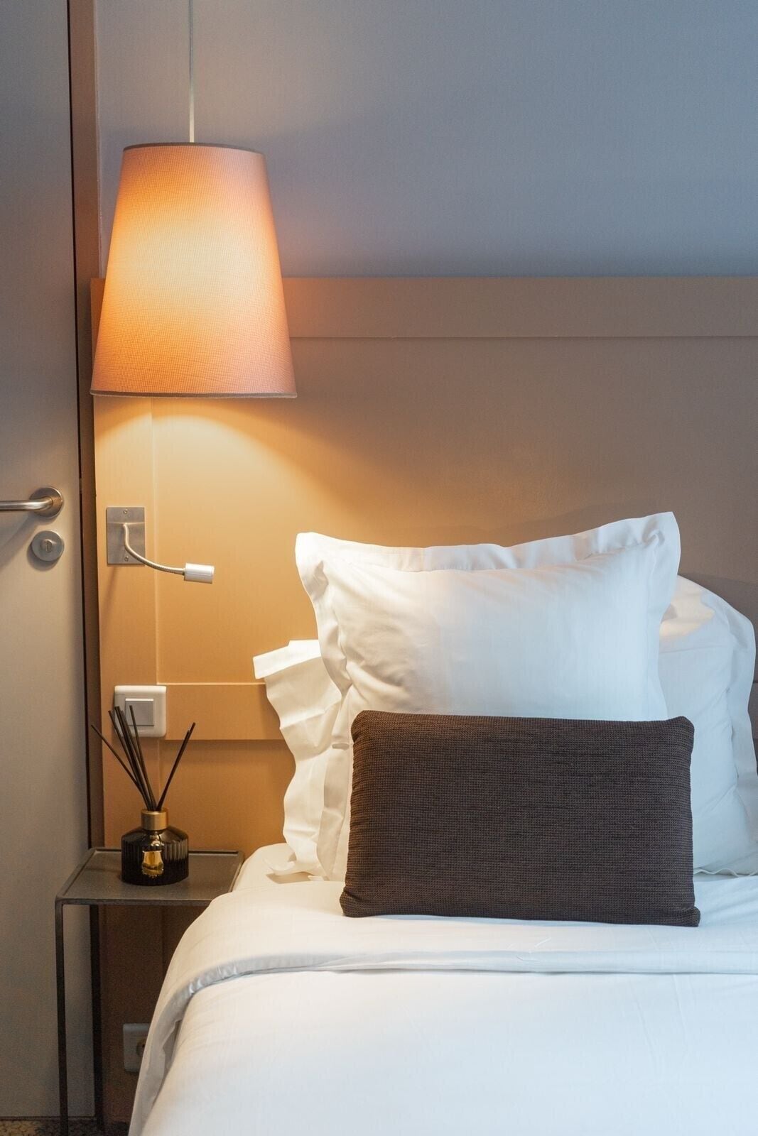 A neatly made bed with white and brown pillows, a bedside table with a lamp, and a wall socket.