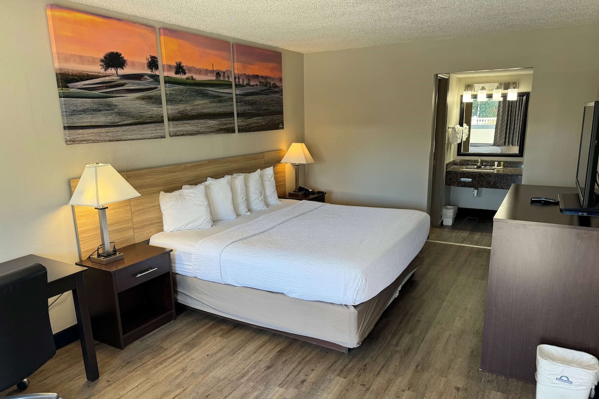 A hotel room with a large bed, two bedside tables with lamps, a wooden headboard, a painting on the wall, a bathroom with a sink and mirror, and a wooden floor.