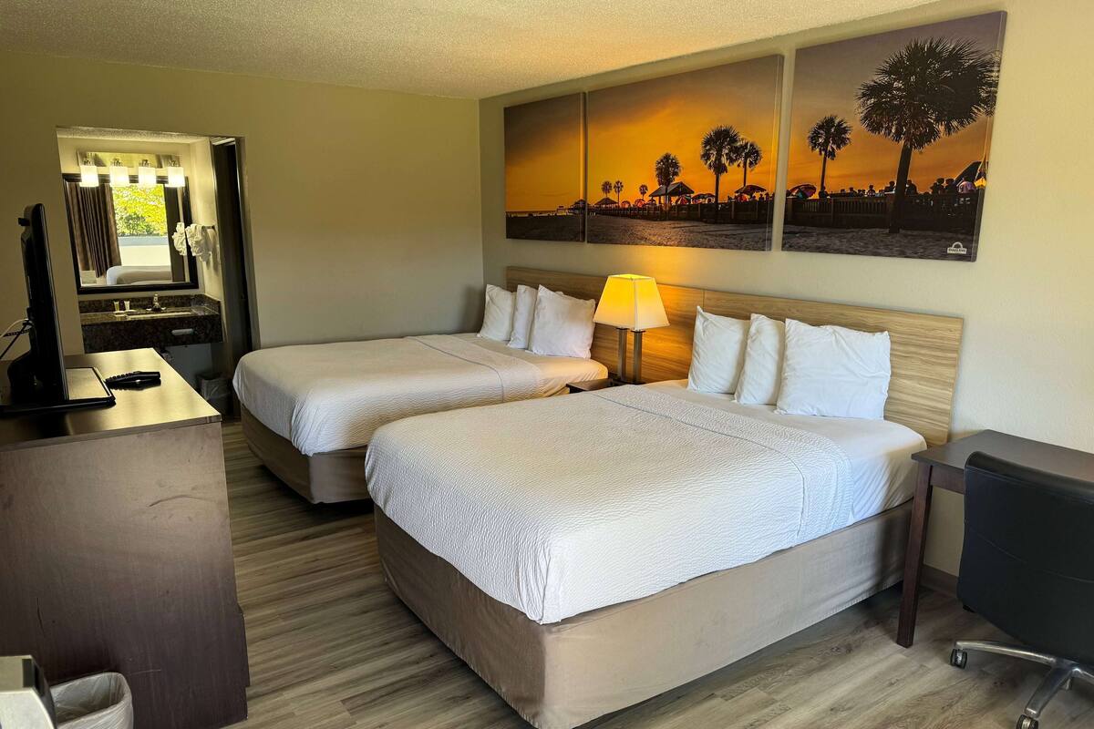 A hotel room with two beds, a desk, a mirror, and a painting of a sunset.