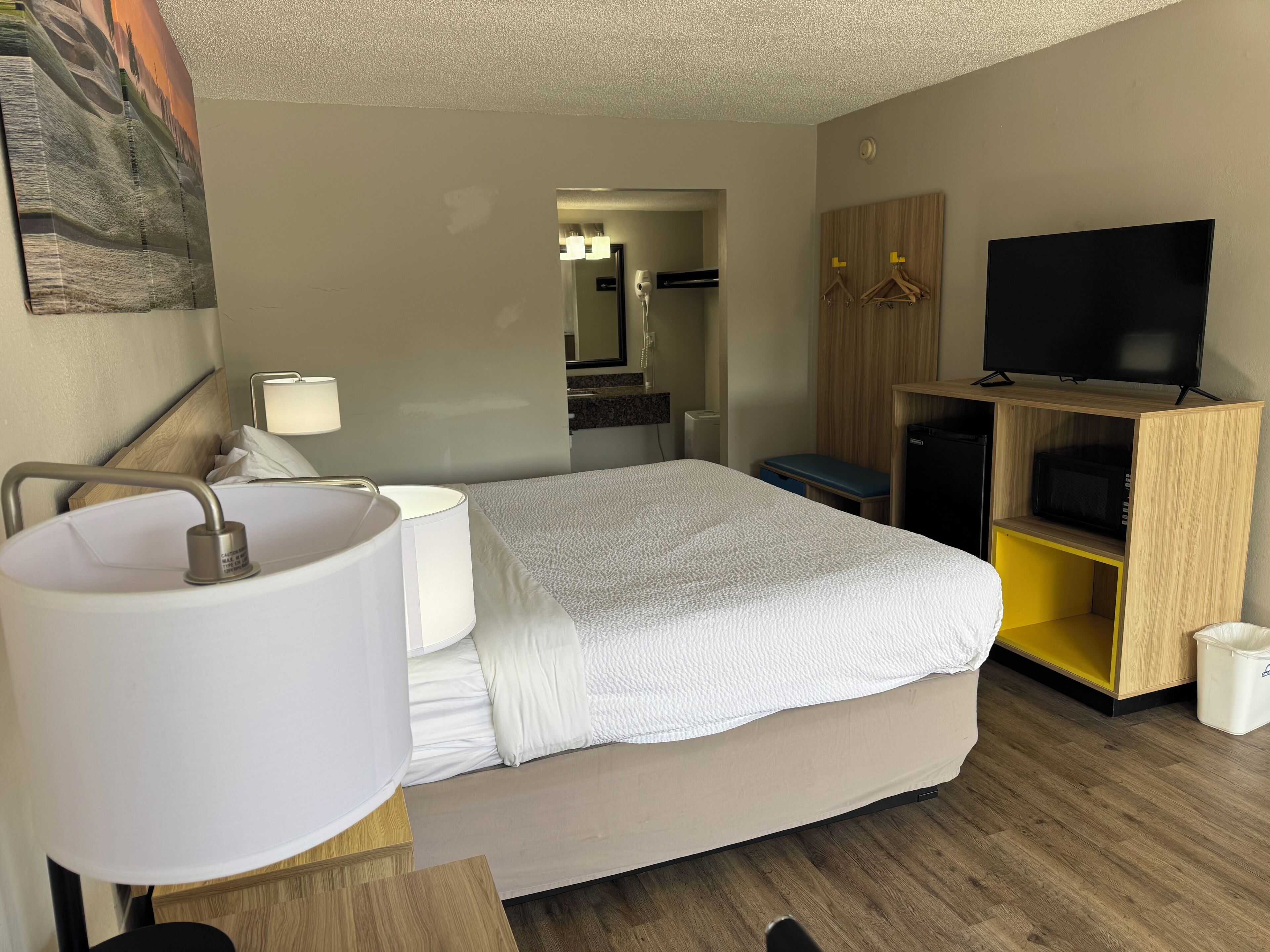 A hotel room with a bed, bedside table, lamp, TV, wardrobe, and a bathroom visible in the background.