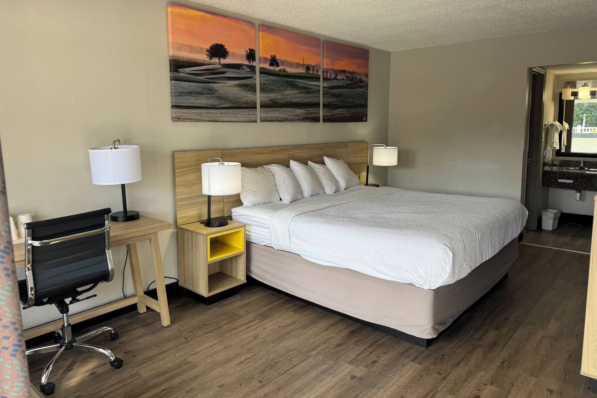 A hotel room with a bed, bedside lamps, a desk with a chair, a small yellow shelf, and a painting on the wall.