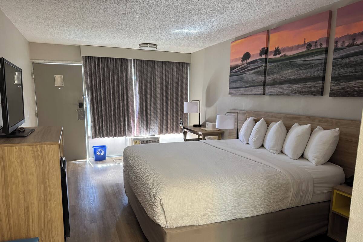 A hotel room with a large bed, a desk with a computer, a TV, and a painting of a sunset on the wall.