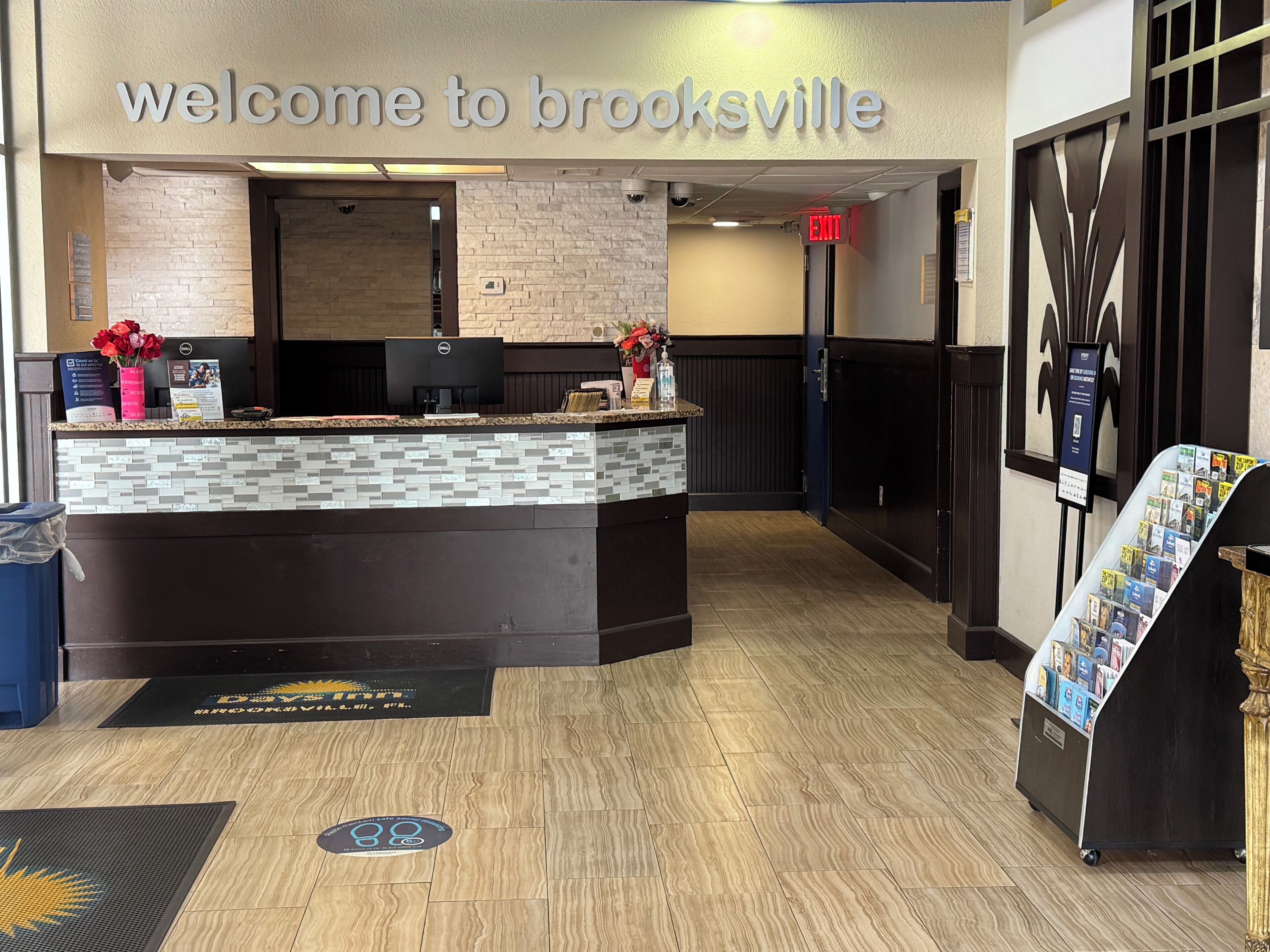 Un área de recepción con un cartel que dice 'Welcome to Brooksville', un mostrador de recepción, una computadora y un tablón de anuncios con folletos.