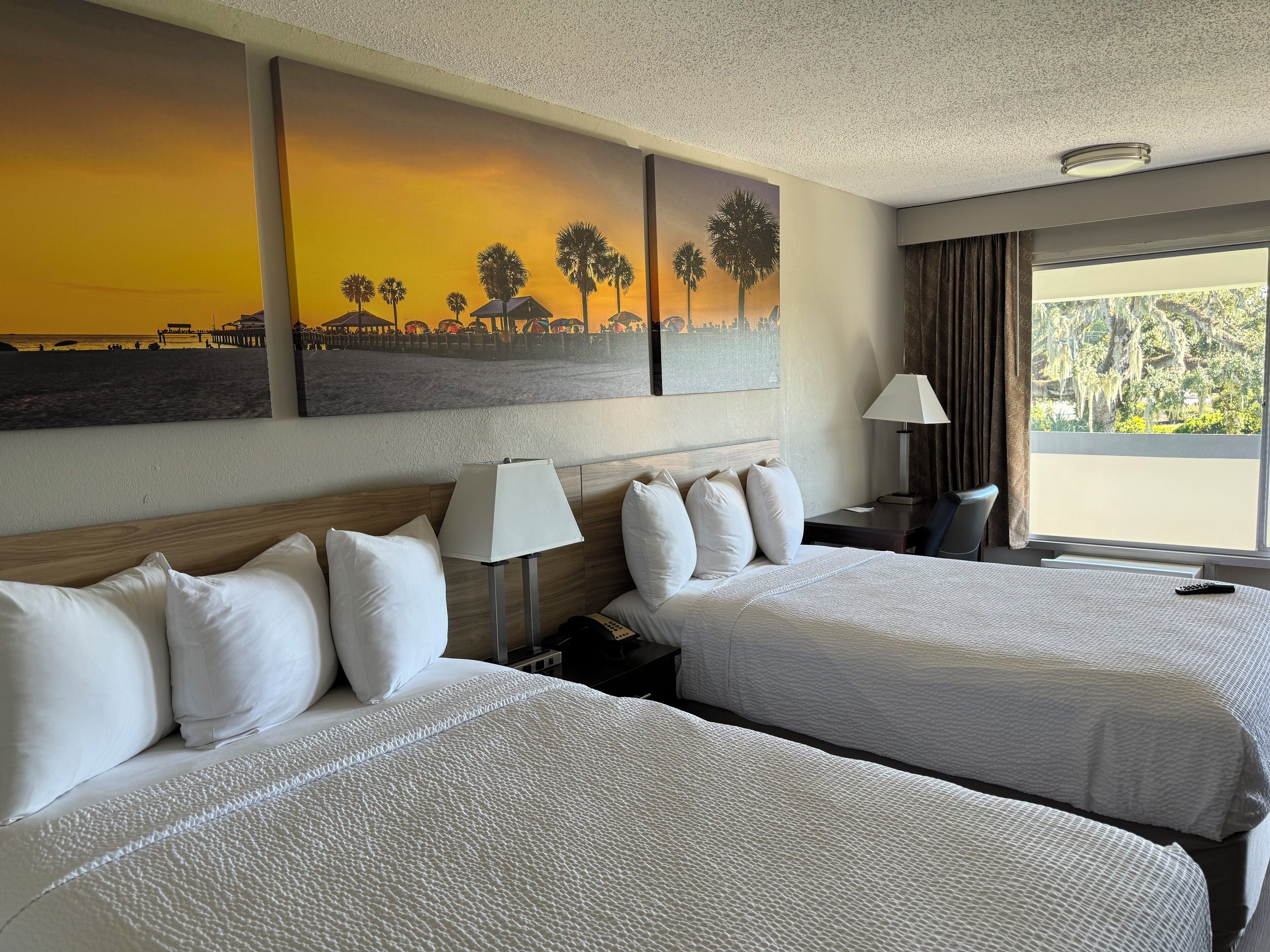 A hotel room with two beds, a nightstand, a lamp, and a painting of a sunset beach scene.