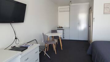 A compact hotel room with a bed, a desk with a chair, a small kitchenette, and a TV mounted on the wall.