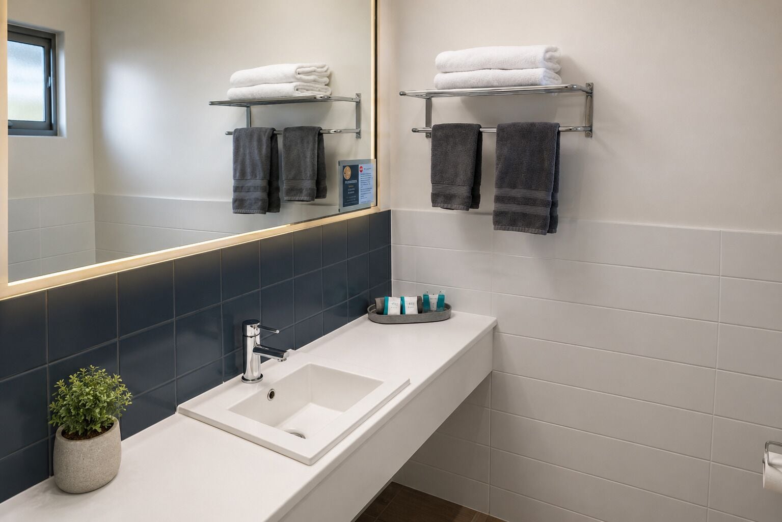Un baño moderno con un lavamanos blanco, salpicadero de azulejos oscuros, un espejo y dos toalleros con toallas dobladas.