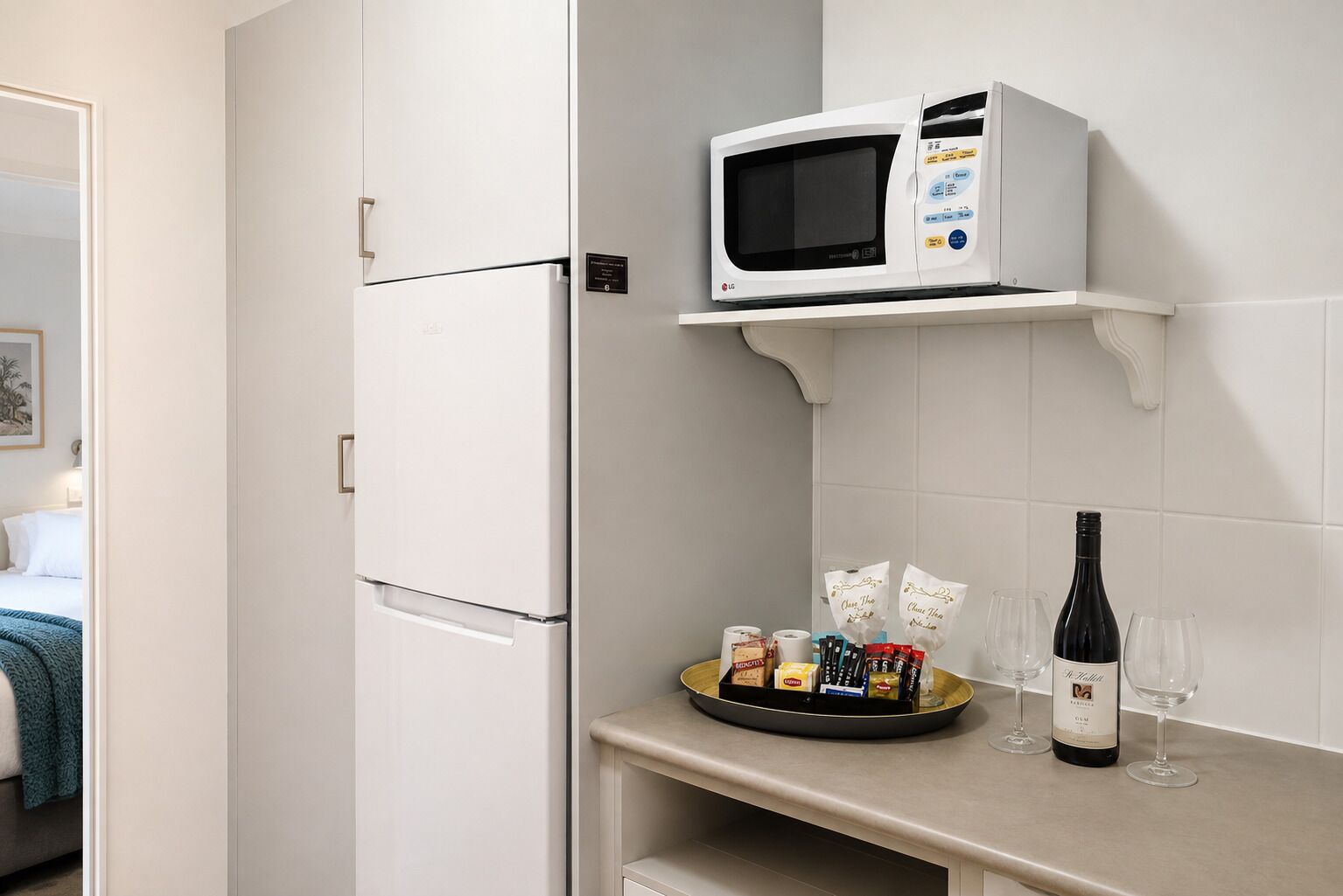 Une chambre d’hôtel avec un réfrigérateur blanc, un four à micro-ondes posé sur une étagère, un plateau avec des en-cas et une bouteille de vin, et un lit recouvert d’une couverture bleue.