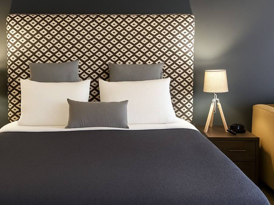 Un lit bien fait, avec une tête de lit à motifs, deux tables de chevet avec des lampes et une chaise, dans une chambre d’hôtel moderne.