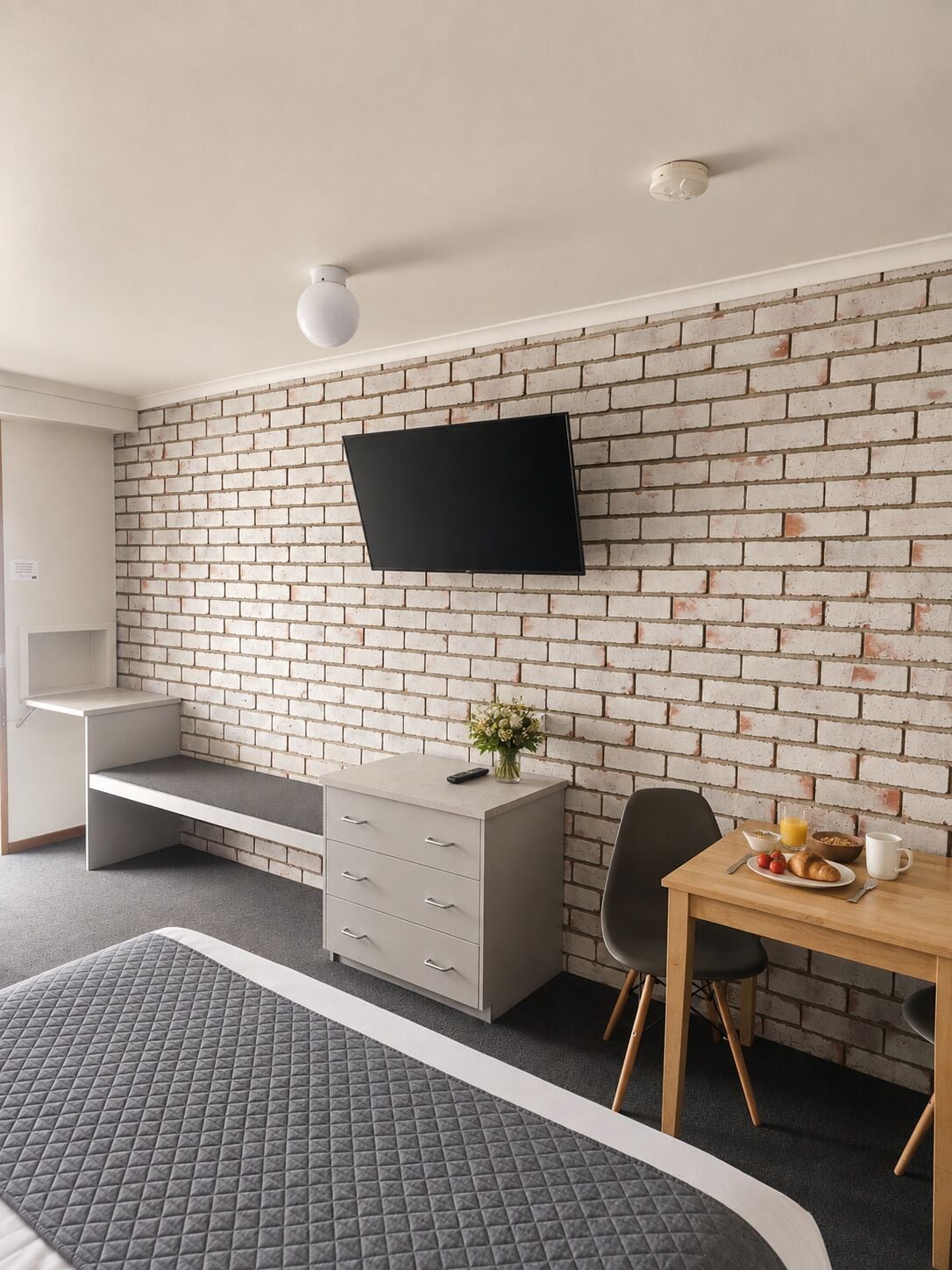 A modern room with a brick wall, a flat-screen TV mounted above a dresser, a bed with a quilted cover, and a breakfast table with a bowl of cereal and a cup.