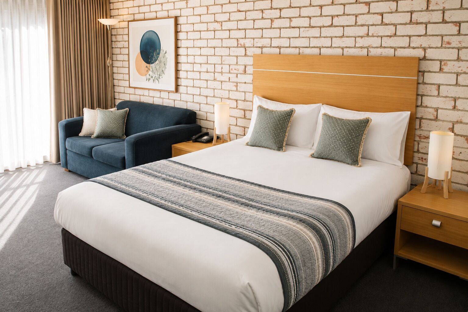 Habitación de hotel con una cama grande, un sillón azul, una mesita de noche con lámpara y una pared de ladrillo.
