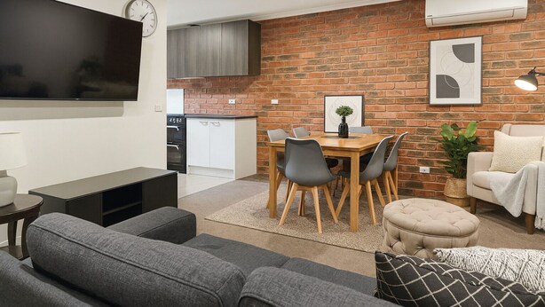 A modern living room with a grey sofa set, a wooden dining table with chairs, a TV mounted on the wall, and a brick wall with a framed picture.