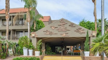 Entrada de hotel con fachada de piedra, palmeras y techo de tejas rojas.