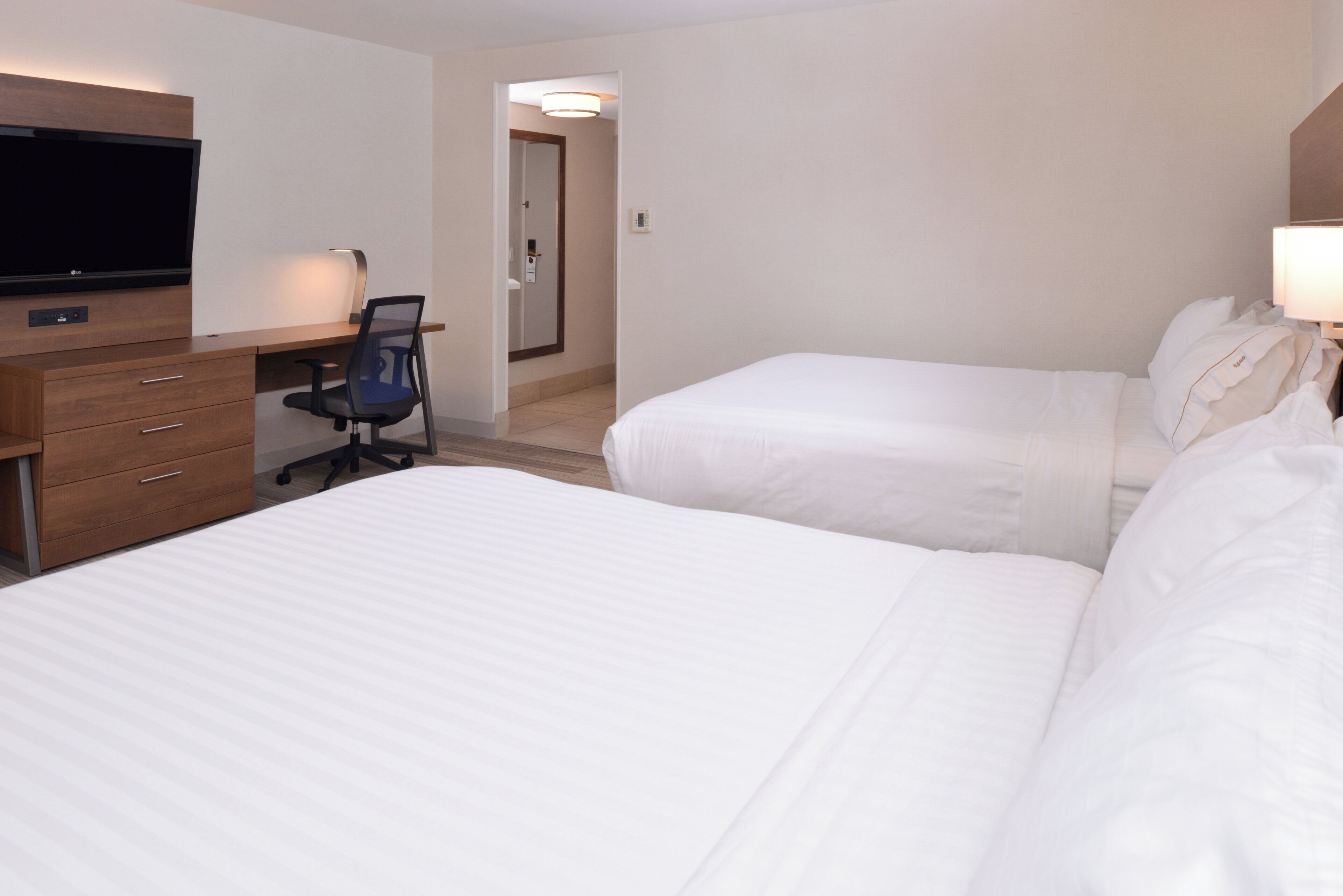 Quarto de hotel com duas camas, uma escrivaninha com cadeira, televisão e uma porta que dá para outro cômodo.