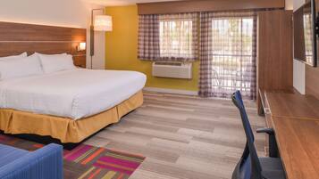 A hotel room with a bed, desk, chair, air conditioning unit, and a window with curtains.