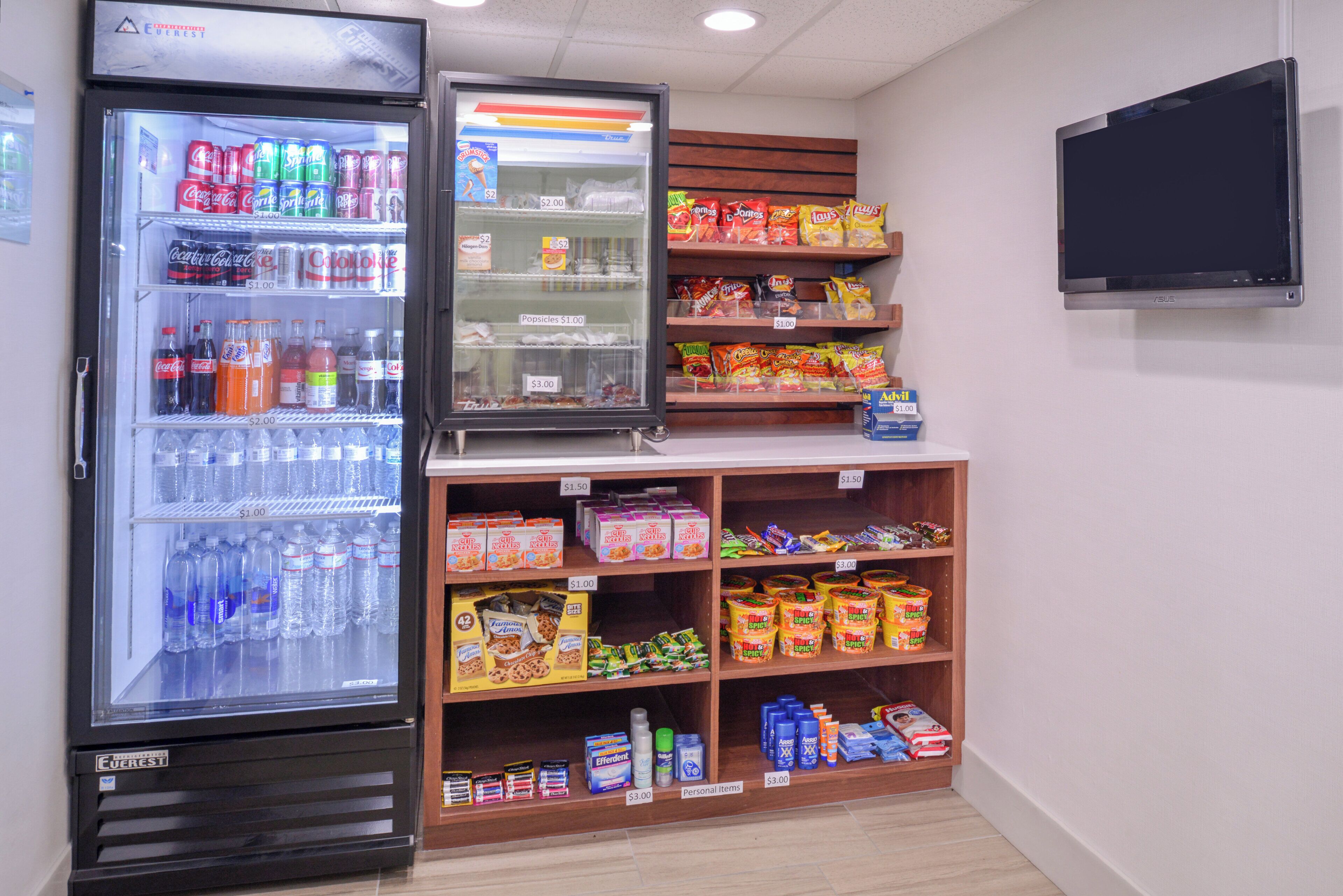 Máquina expendedora con diversas bebidas y aperitivos, un refrigerador con bebidas, y estantes llenos de bocadillos y artículos personales.