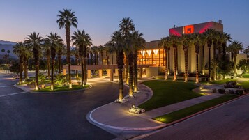 Entrada de un hotel con palmeras, un camino curvo y un edificio con ventanas iluminadas.